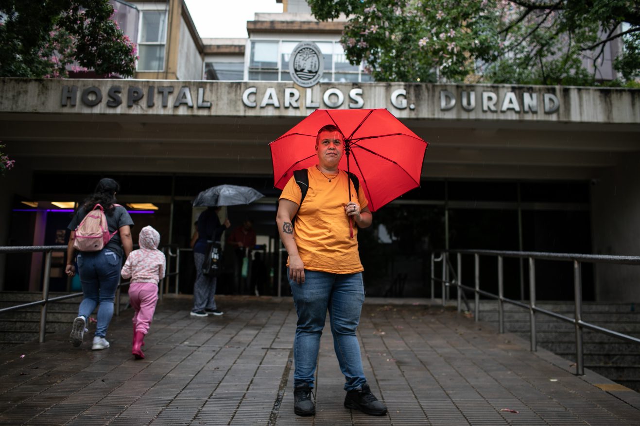 Tom Mascolo standing in front of Hospital Carlos C. Durand wearing jeans, an orange shirt, and holding a red umbrella.
