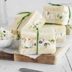 Stacked chicken salad sandwiches with crusts removed on a wooden board, garnished with fresh herbs.