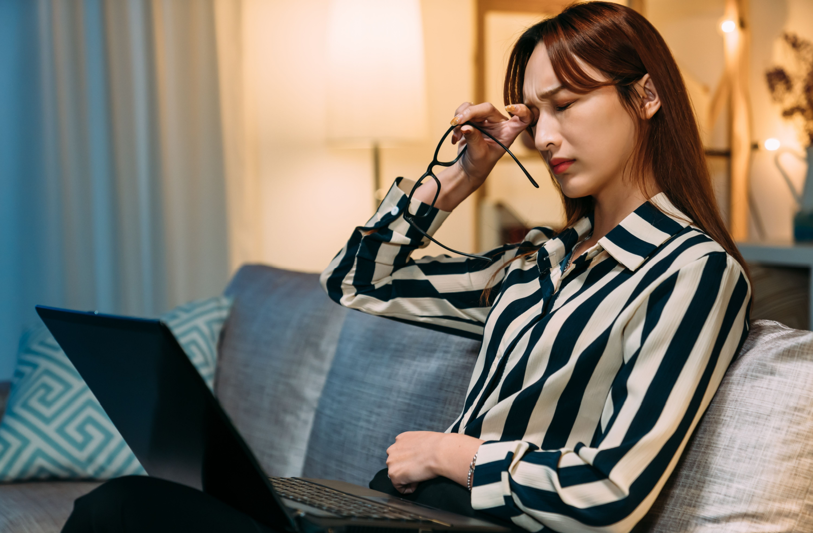 A young person sits on their couch with a computer in their lap, rubbing their eye and holding their glasses.