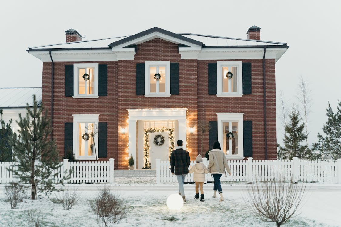Family of three walking toward a brick house in winter, with a front yard featuring dry plants and a light covering of snow.