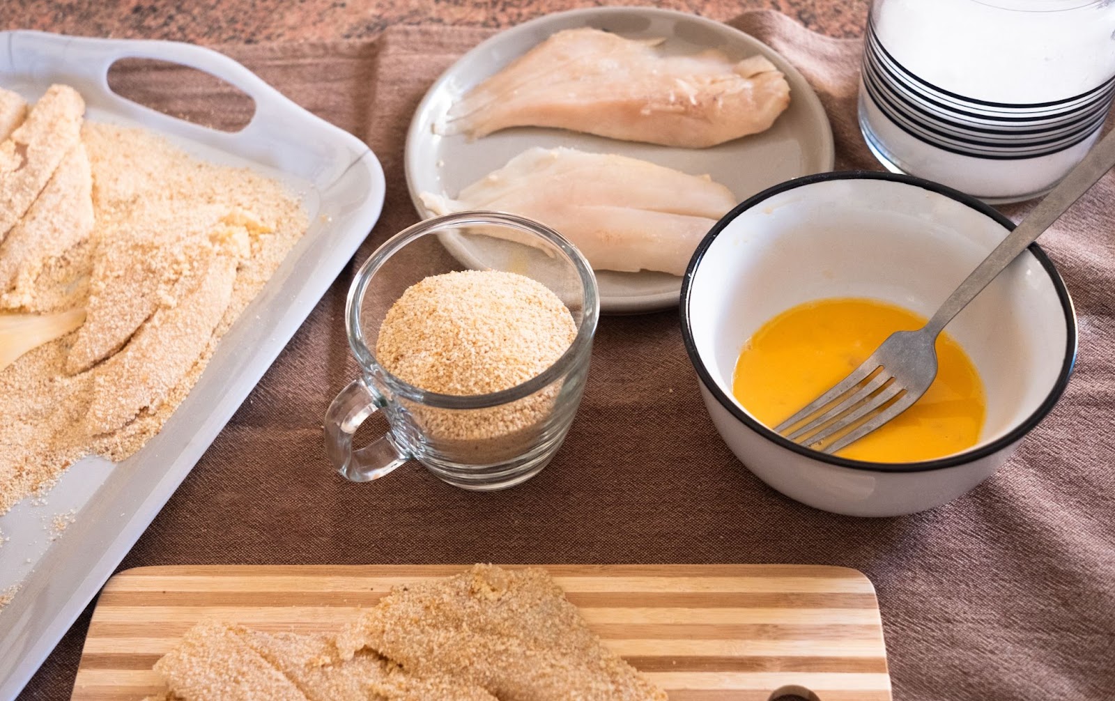 a breading station with chicken cutlets fully coated with egg and bread crumbs resting in a tray and wooden board