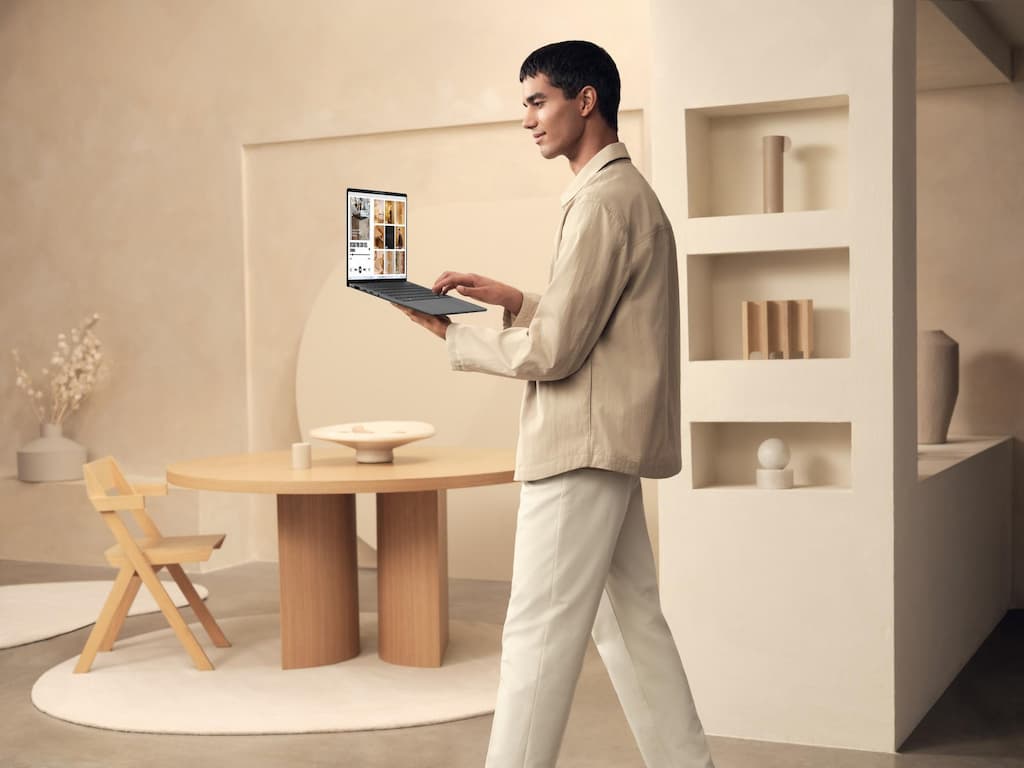 Um homem, com camisa bege e calça branca, caminhando enquanto trabalha em seu Zenbook A14, o ambiente ao fundo é moderno e em tons claros. 