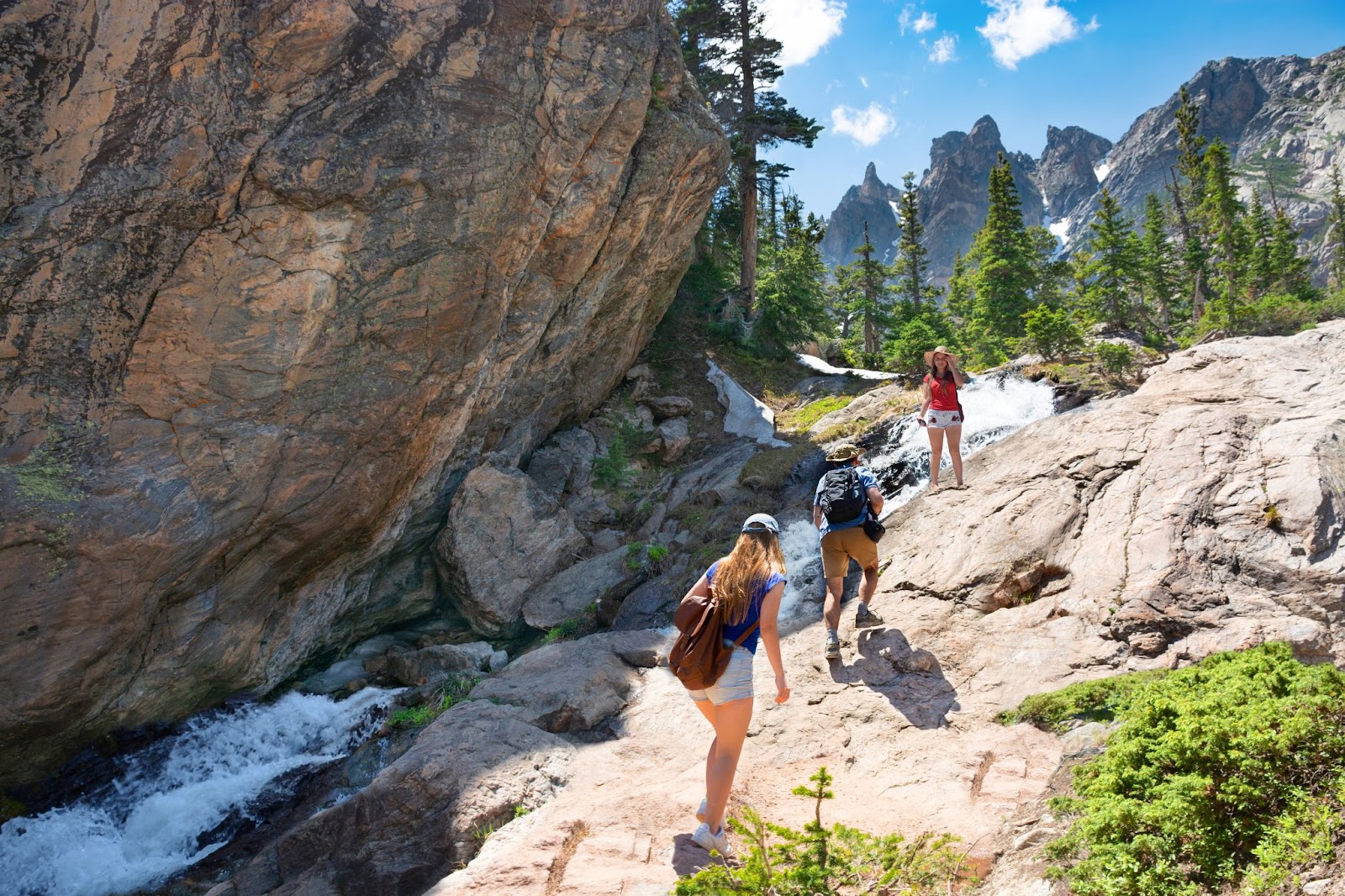 Three girls climb over rocky terrain next toa small waterfall, a tall rock cliff next to them, jagged peaks visible in the background