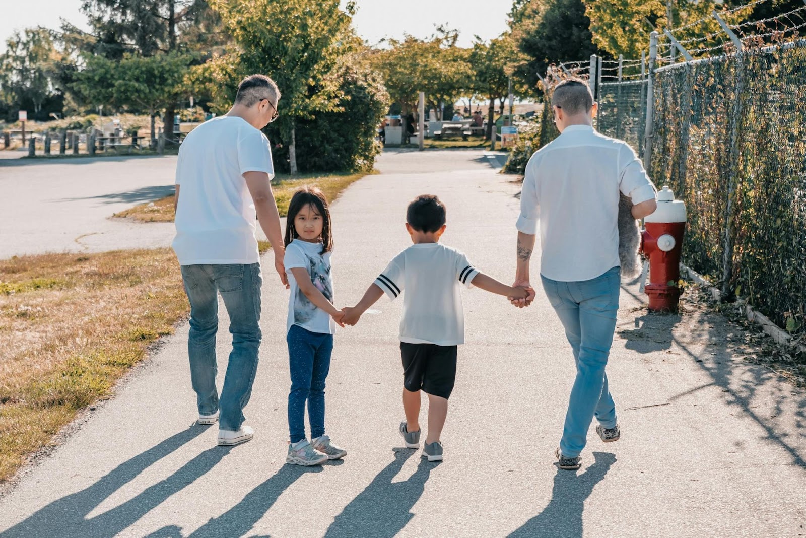 "Two adults walking hand-in-hand with two young children on a sunny pathway, surrounded by greenery and a fence on one side. The girl looks back at the camera, creating a warm family moment."