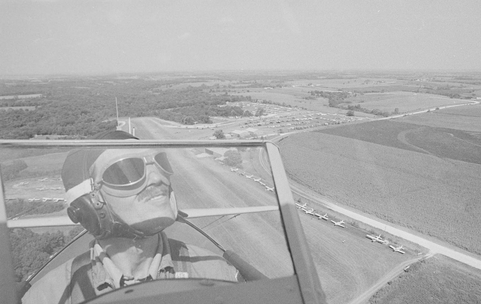 A black & white photo of a pilot wearing helmet and goggles in an open-air cockpit, frying over an airport runway and surrounding area with trees and fields