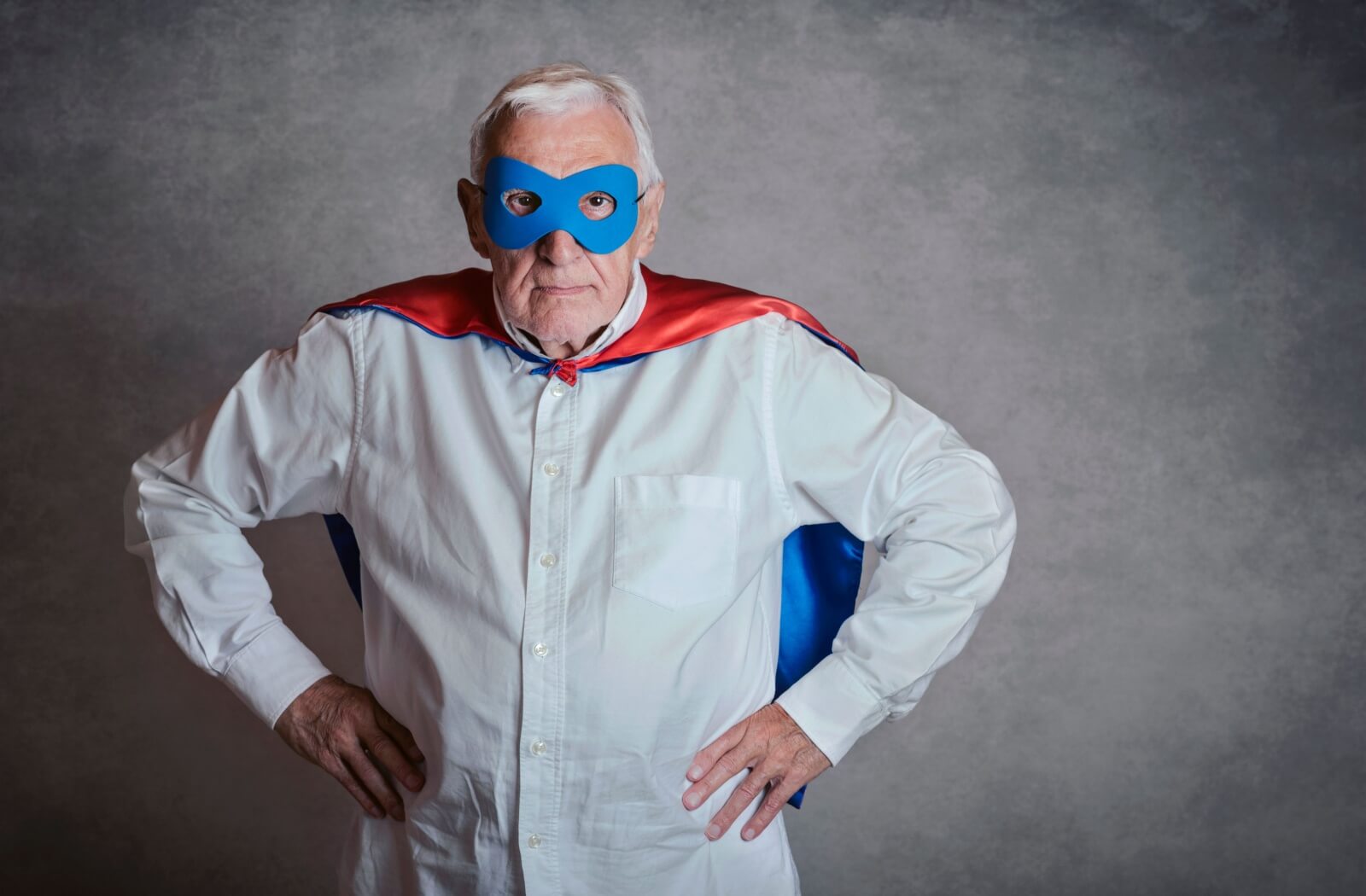 A senior with hands on their hips dressed up as a serious-looking superhero with a blue mask and blue and red cape.