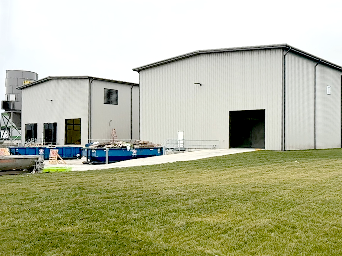 Two large metal industrial buildings with an open entrance on one, surrounded by green grass and construction materials, including blue dumpsters and a silo in the background.