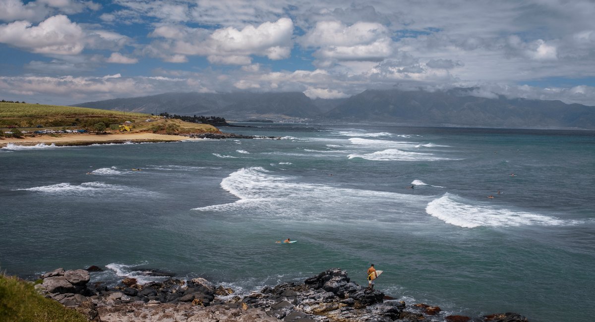 Scenic view of surfers riding waves on a vibrant day at Hookipa Beach beach in Maui, Hawaii, with rocky coastline in the foreground and dramatic mountain backdrop under a partly cloudy sky.