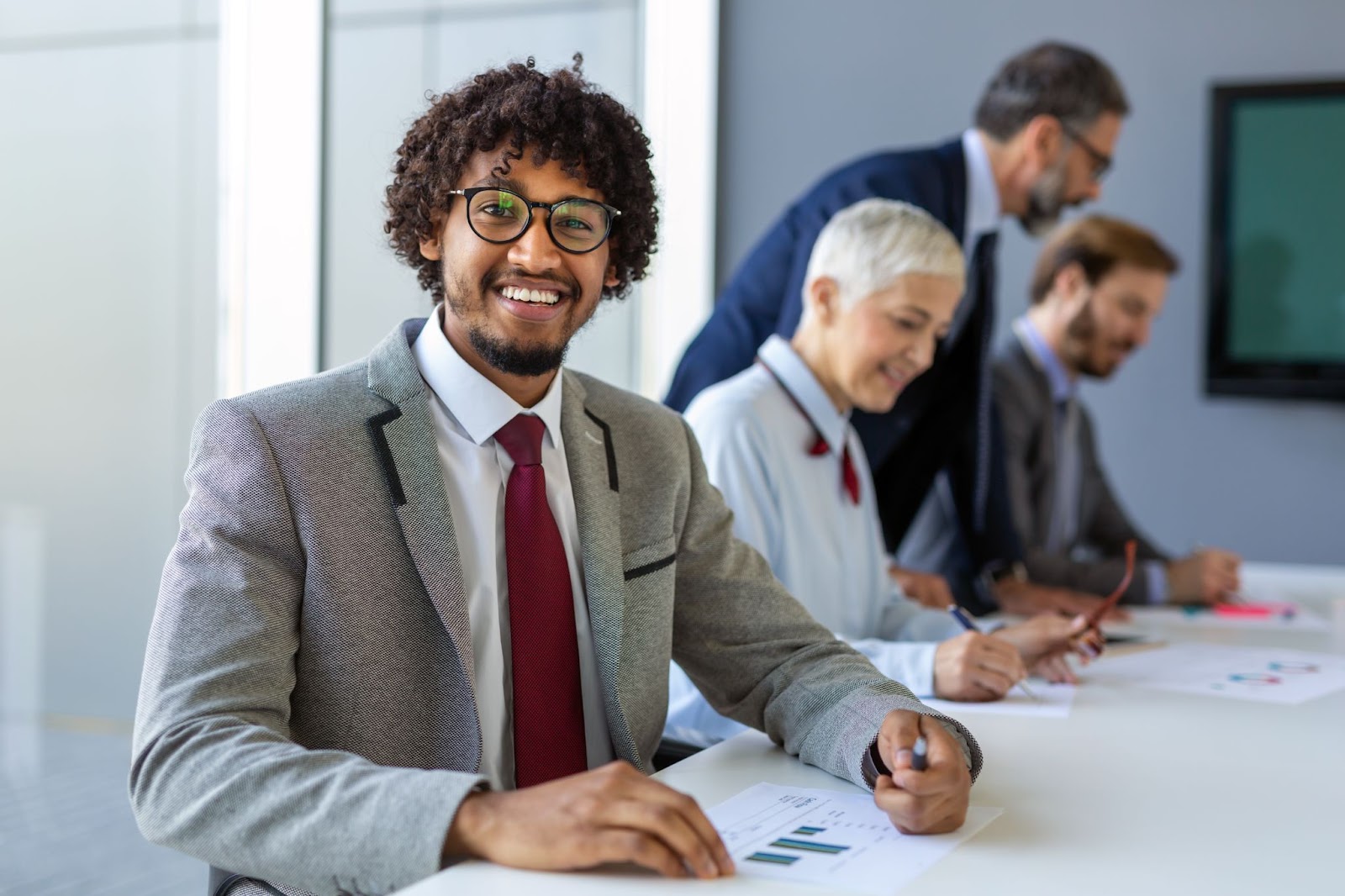 Three business professionals in business attire look at documents in an office, and another business professional wearing glasses, a white collar shirt, a red tie, and a gray jacket smiles into the camera.