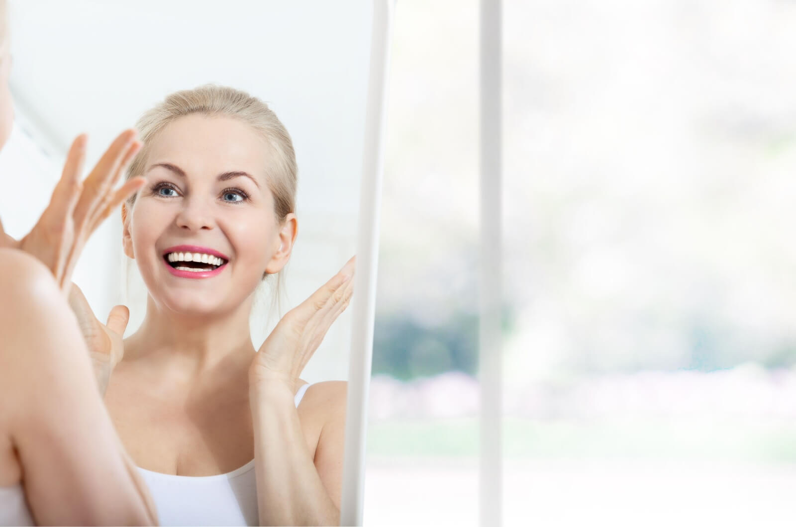 A smiling middle-aged woman lifting her hands in surprise while looking at her face in the mirror after getting Botox.