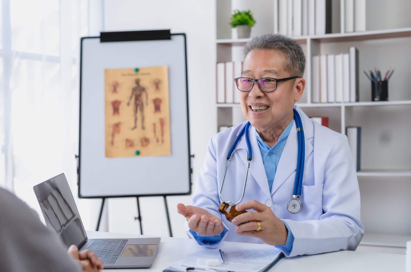 A senior male doctor in a lab coat with a stethoscope around his neck discusses treatment options with an unseen patient. A human anatomy chart on an easel and white bookshelves are in the background.