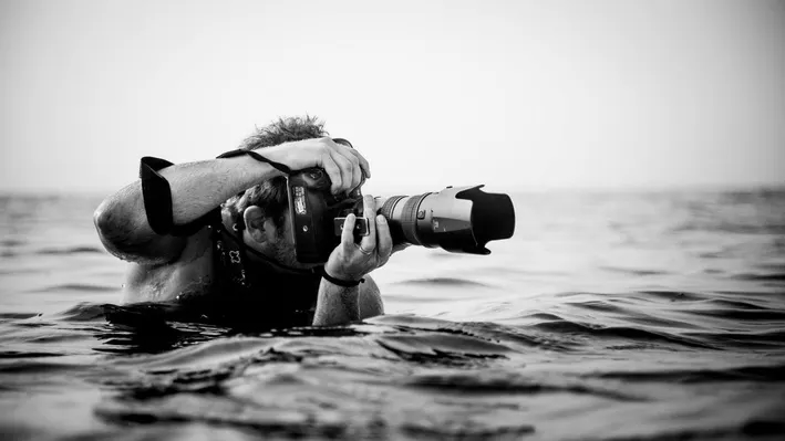 Chase Jarvis standing in the ocean with the water up to his chest while taking photos with a large camera.