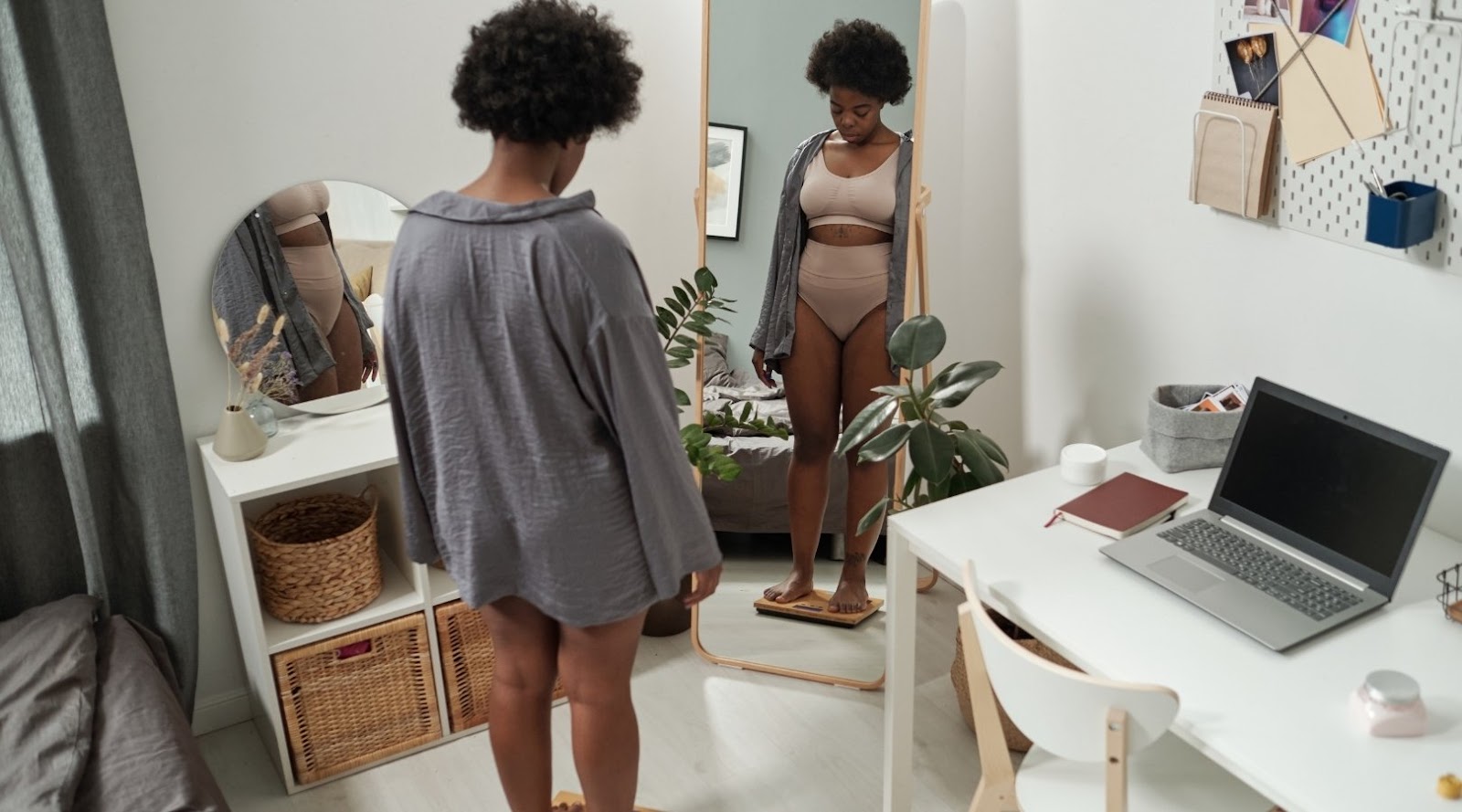 Femme debout sur une balance devant un miroir, portant une robe de chambre et des sous-vêtements, vérifiant son reflet dans une chambre confortable.