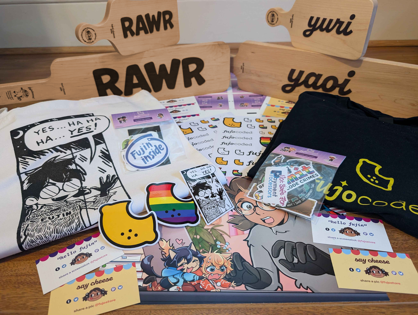 An array of FujoCoded merchandise on a wooden floor. In the back of the array are four fujoboards, with a chibi version perching on top of the two larger hime versions, all standing on end so the inscribed text is visible. Two say “rawr” and the other two say “yuri” and “yaoi.” Underneath the other merchandise are several sheets of FujoCoded logo stickers and a print of Boba-tan watching Git and GitHub squabble. On the left and right of the pile are two folded tee shirts. On top of the tee shirts are sticker packs and large FujoCoded lemon logo stickers. At the front of the array are four packing cards with FujoCoded branding that say either “hello fujin” or “say cheese.”