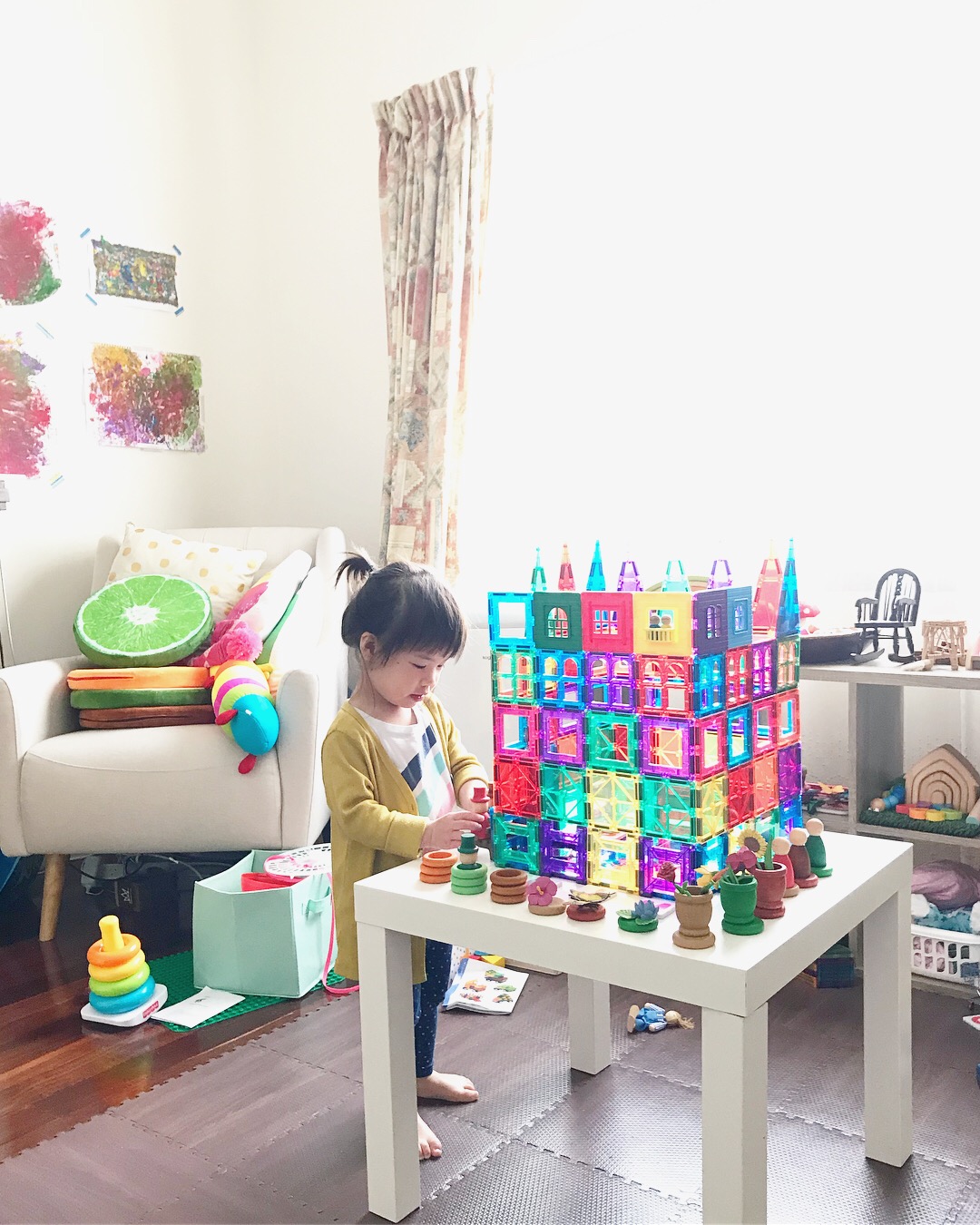 A child is building a tall castle using colorful magnetic tiles, adding loose parts like rings, cups, coins, and peg dolls around the structure. Through the process, the child is developing important life skills such as resilience, perseverance, and mental strength to face setbacks while creatively exploring and problem-solving.
