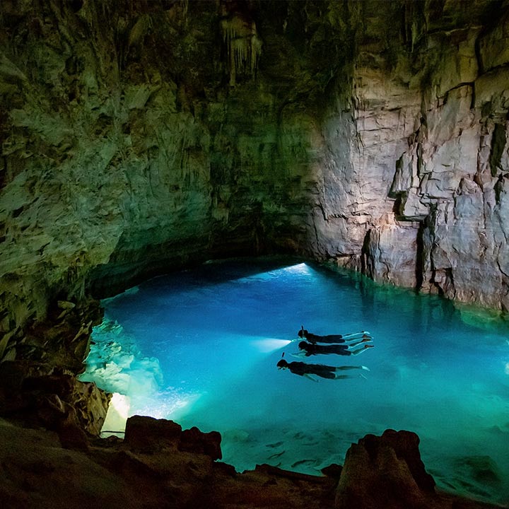 Turistas mergulhando em uma caverna com água azul cristalina no interior, rodeada por paredes rochosas e vegetação na entrada da caverna