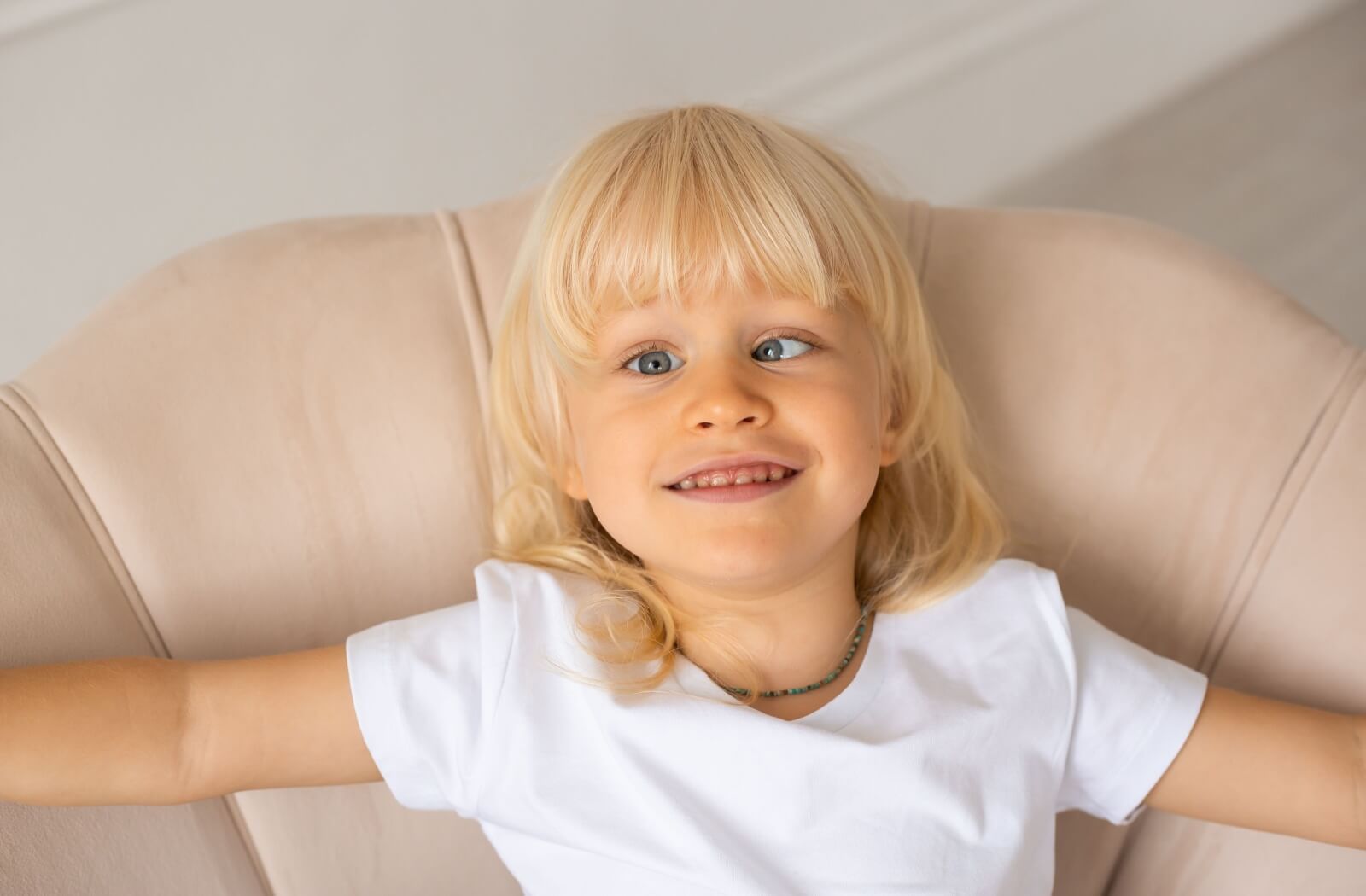 Smiling toddler sitting in a beige chair, with visible signs of strabismus, highlighting a relaxed and cheerful demeanor.