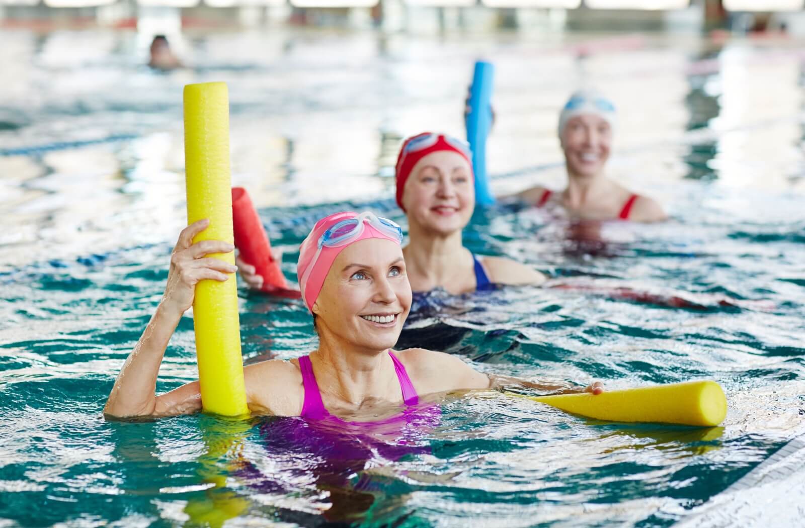 Three senior woman hold pool noodles underneath them in a pool, smiling at an offscreen water aerobics instructor