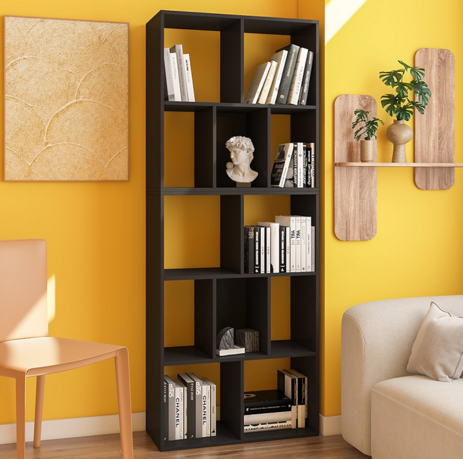 A modern living space featuring home furniture, including a tall black bookshelf with an open-cube design, holding books and decorative items against a vibrant yellow wall. The room also includes a beige chair and a matching sofa, complemented by wall-mounted wooden shelves and minimalist decor, creating a warm and inviting atmosphere.
