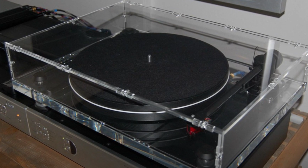 A turntable with a transparent acrylic dust cover, resting on an audio system with a felt mat on the platter.