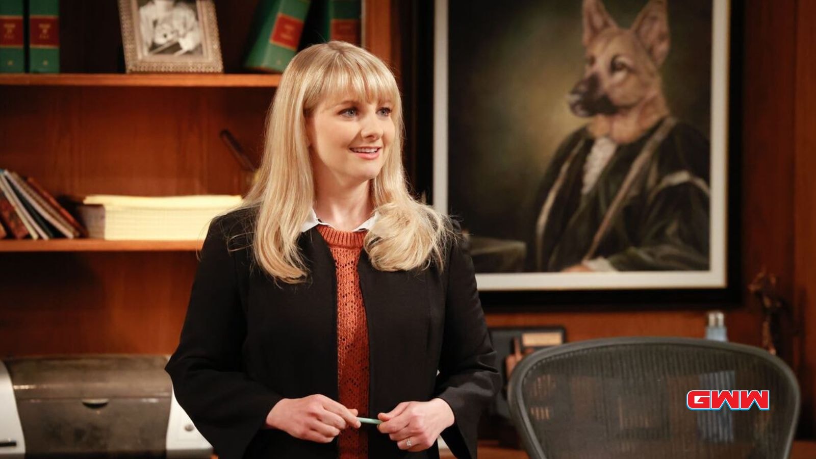 Abby Stone in black blazer smiles with portrait of dog behind her.