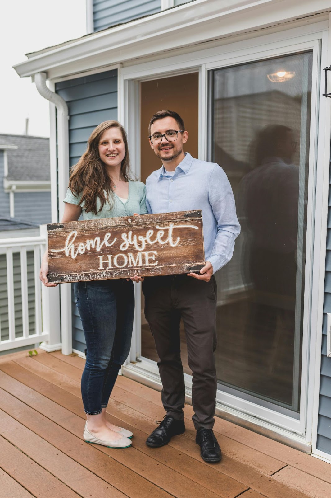 Smiling couple standing in front of their new home, holding a wooden sign that reads 'Home Sweet Home,' symbolizing homeownership and a fresh start. This is their first mortgage.