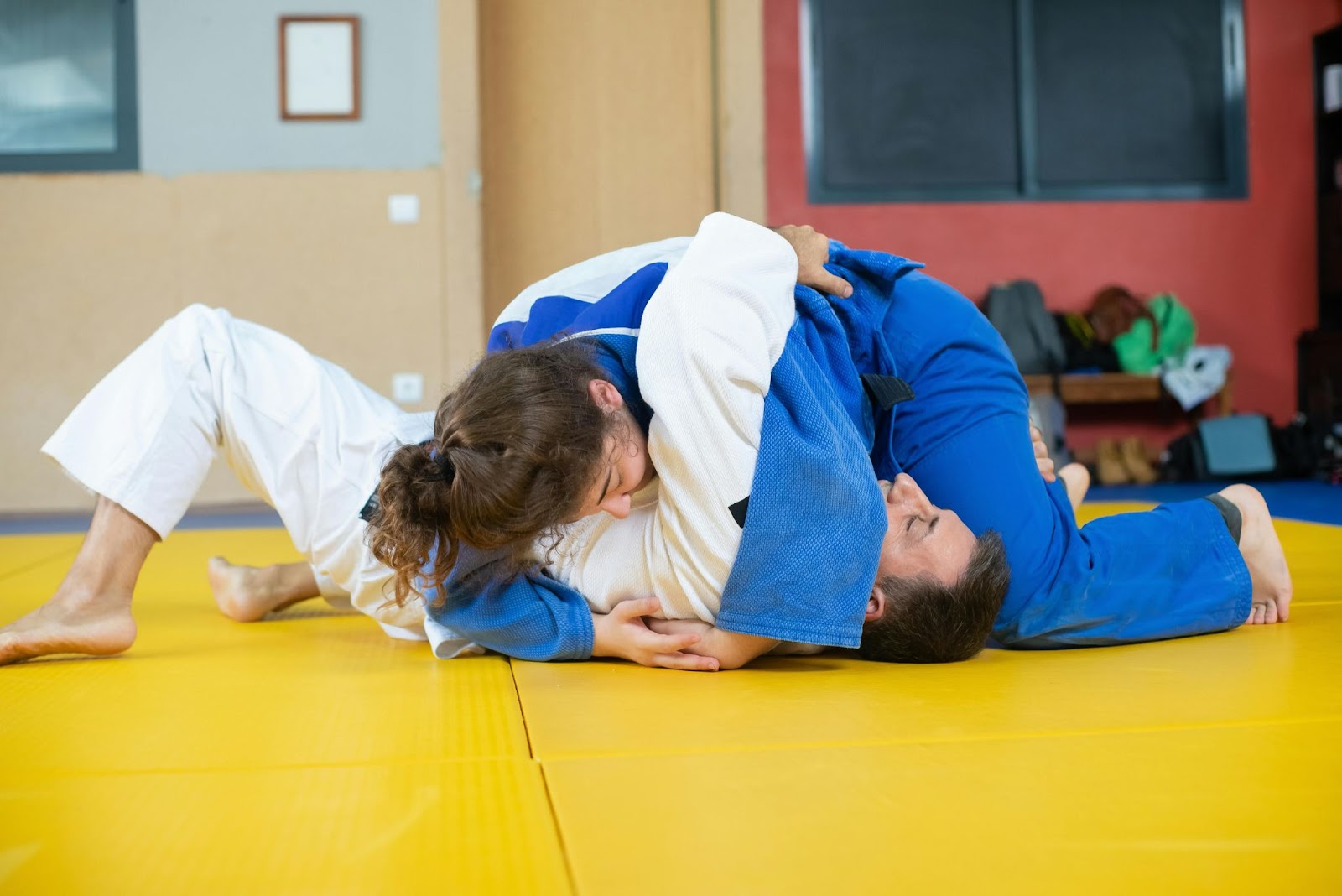 Two young martial arts students engaged in floor grappling, wearing training uniforms in a brightly lit dojo.






