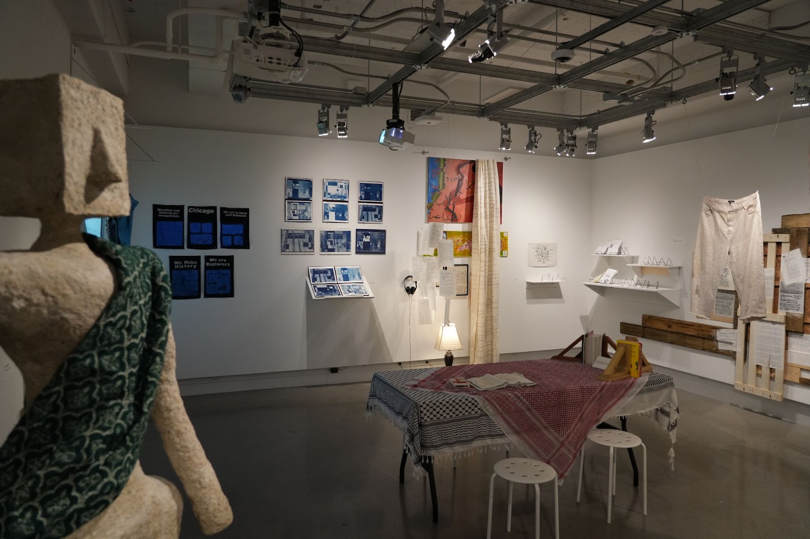 Image: Installation view of School as a Function of Empire. One the left side of the image stands a statue wrapped in a green garment. In the background, other pieces of the exhibition including photos, maps, and letters can be seen. Photograph by Curators Under Censorship. 