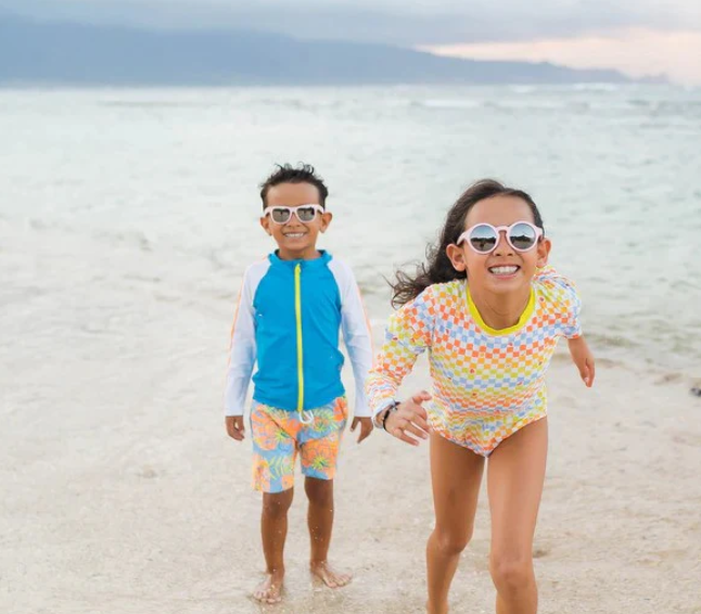 Brother and sister kids at the beach smiling with polarized sunglasses, wearing sun-protective swimwear by SwimZip—10 ways to keep your kids safe during summer vacation