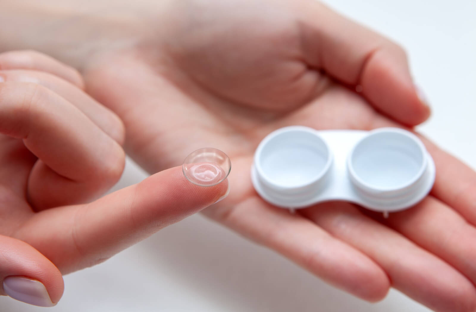 A close-up image of a contact lens case in one hand with the lens on the tip of a person's outstretched index finger.