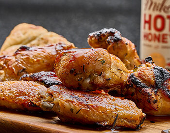 A plate of golden-brown grilled chicken wings glazed with hot honey, served on a wooden board with a side of bread and a bottle of hot honey.