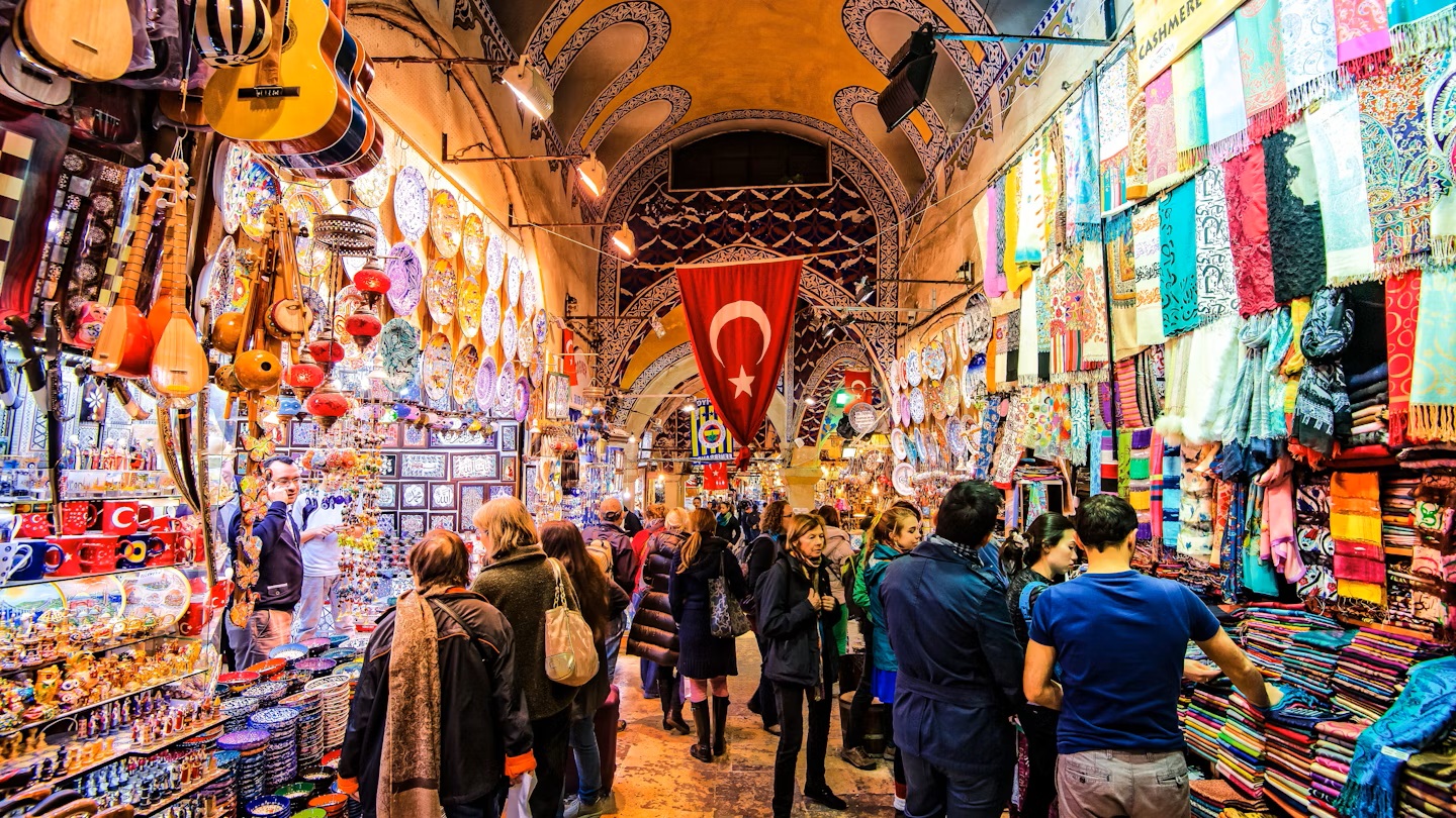 Colorful interior of the Grand Bazaar in Istanbul, Turkey, with crowds shopping for spices, textiles, souvenirs, and traditional crafts under ornate ceilings and Turkish flags.