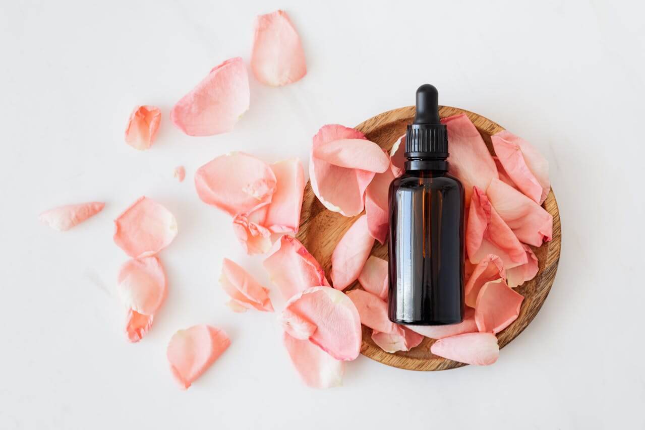 Pink rose petals on a wooden plate beside a bottle of essential oil, highlighting the benefits of scalp massage with herbal oils.