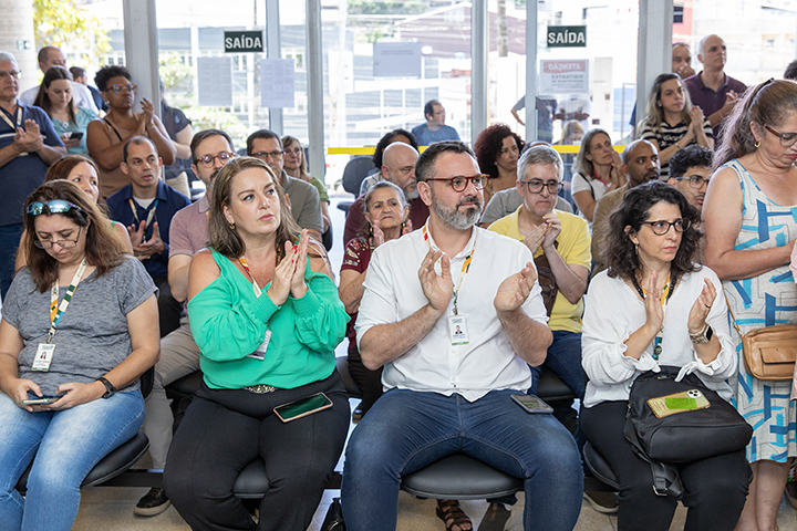 A imagem mostra um grupo de pessoas em um evento ou reunião, provavelmente em um ambiente interno como uma sala de espera ou auditório. Na cena, algumas pessoas estão sentadas em cadeiras na frente, enquanto outras estão em pé ao fundo. Muitas delas estão batendo palmas, sugerindo que estão reagindo positivamente a algo, como um discurso, apresentação ou evento especial.