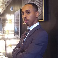 This contains a picture of American streamer Johnny Somali wearing a suit and tie standing in front of a wall with pictures on it