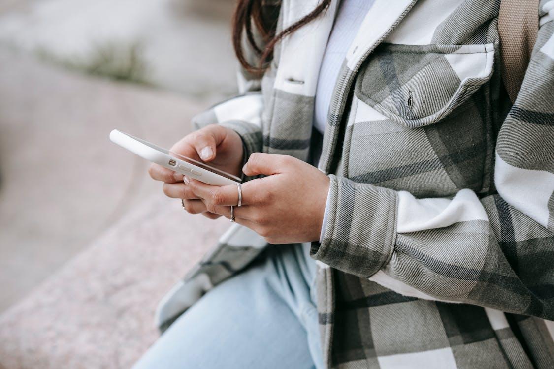 Free Crop anonymous young woman in casual outfit sitting on stone fence and text messaging on phone in daytime Stock Photo