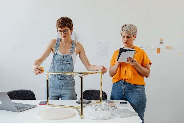 Two people are working at a table in a bright room. One, in overalls, measures the table frame with a tape measure, considering ergonomic solutions. The other, in an orange shirt, takes notes on a pad. A laptop and various materials related to interior design are on the table.