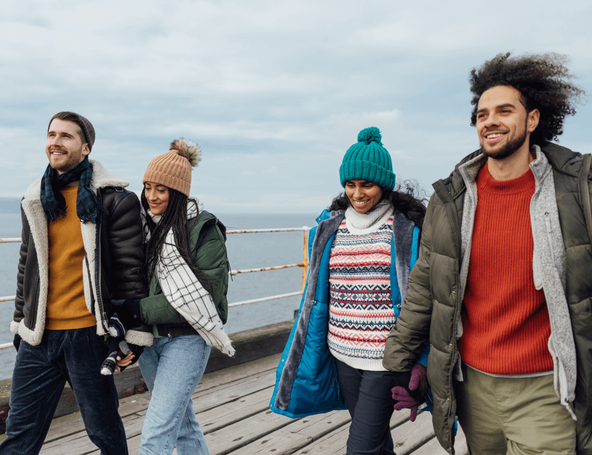 A group of four friends dressed in cozy winter clothing, including jackets, sweaters, and beanies, walking outdoors on a wooden pier.