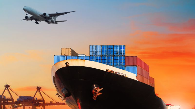 A cargo ship docked at port as an airplane flies overhead at sunset.