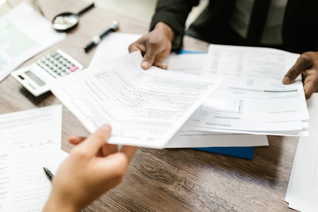 Duas pessoas trocando documentos em uma mesa de trabalho com papéis, calculadora e equipamentos de escritório ao fundo.