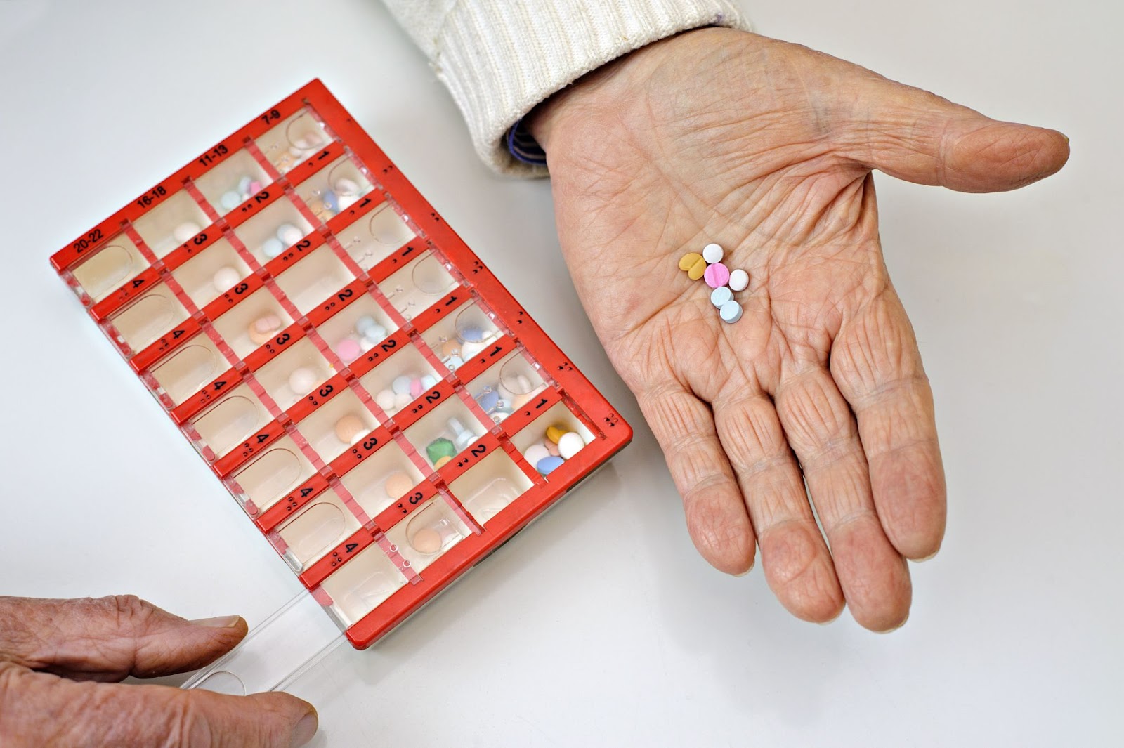 A close up pill organizer on a table with an older adult's hand with medication.