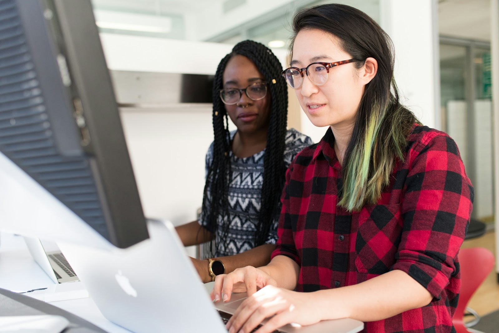 Two women collaborating on a computer in a bright office, focused and engaged in their work together.