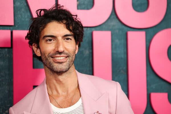 In a shot from the shoulders up, Justin Baldoni smiles against a backdrop with the partial title “It Ends With Us” visible. He’s wearing a pink jacket over a cream-colored top.  