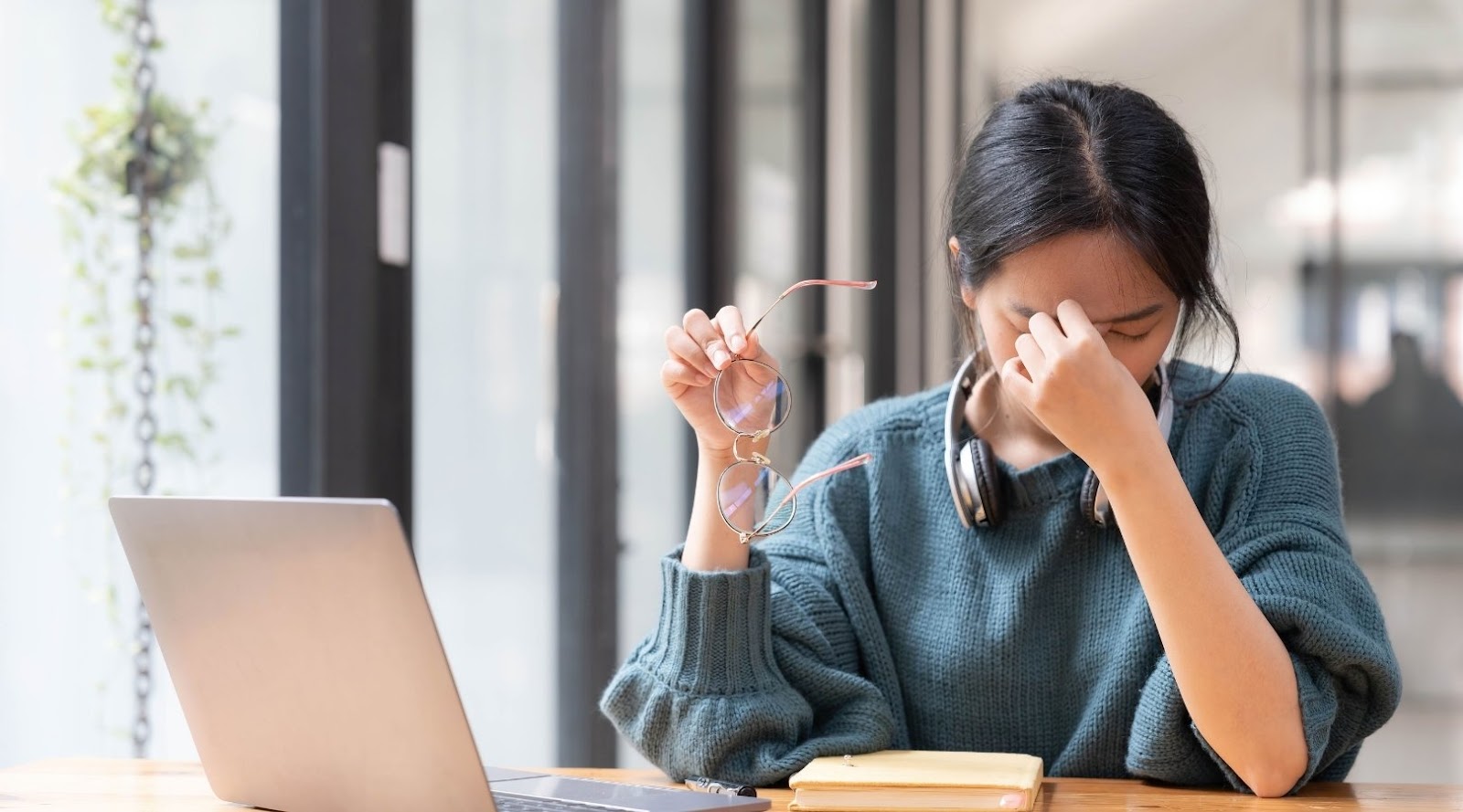 Person holding glasses and rubbing their eyes in frustration, sitting at a desk with a laptop and headphones.