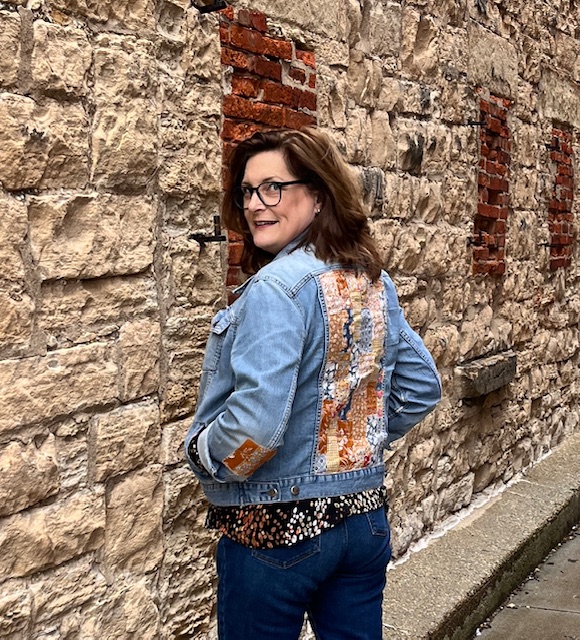 Brunette stands against a brick wall showing the back of her denim jacket with quilted fabric scraps