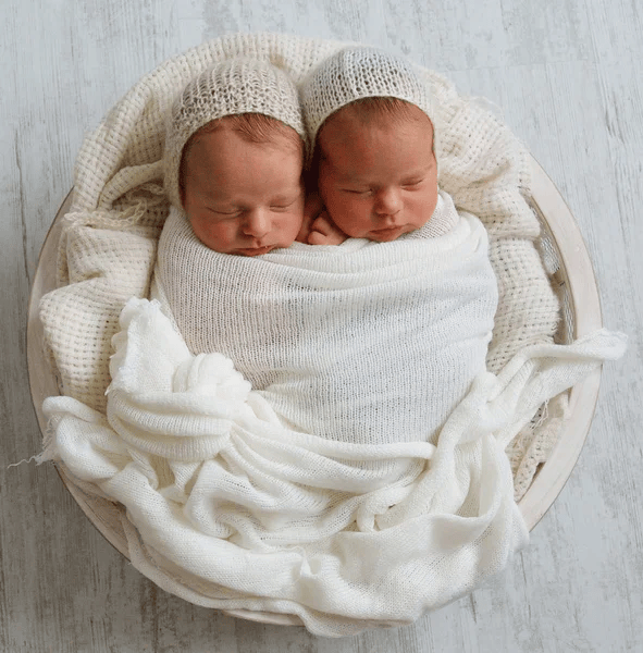 Baby photoshoot featuring twin babies resting in a wooden basket with tangled legs, styled with matching beanies