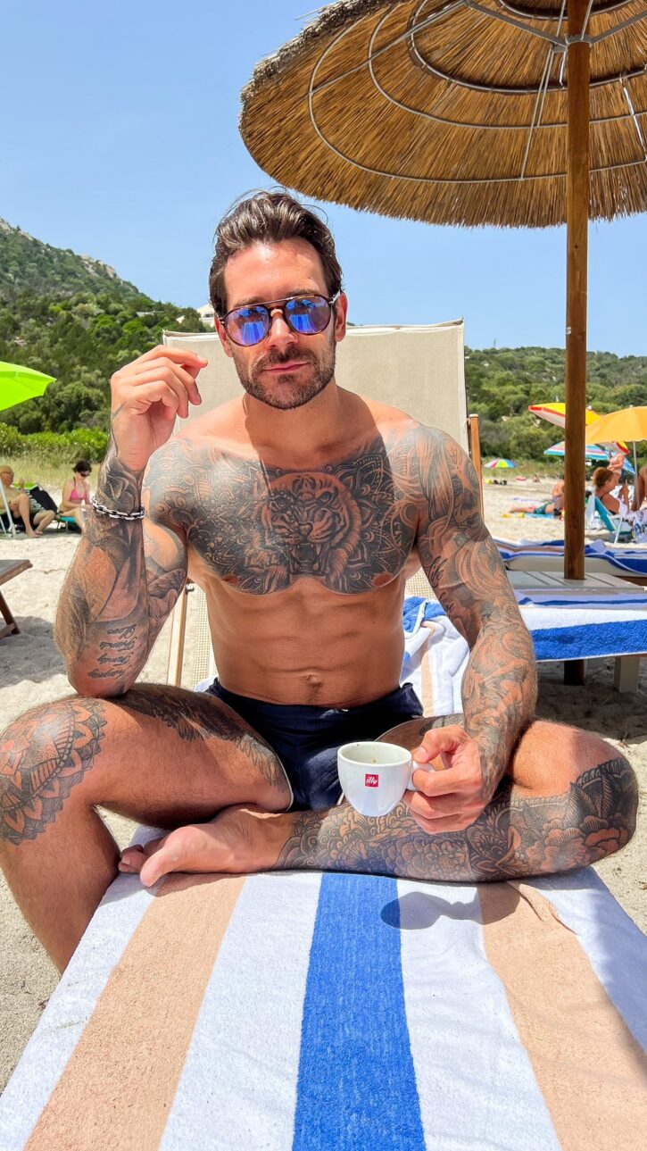 Papi Kocic posing on a beach chair in black speedo with a cup of espresso wearing sunglasses on a striped beach towel