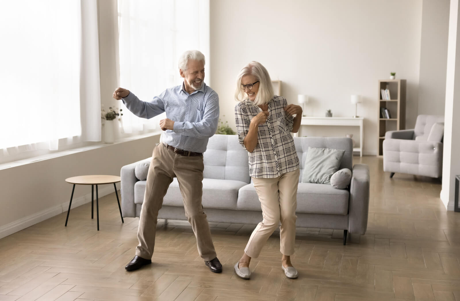 An older couple in a sunlit living room dancing together and laughing with each other.