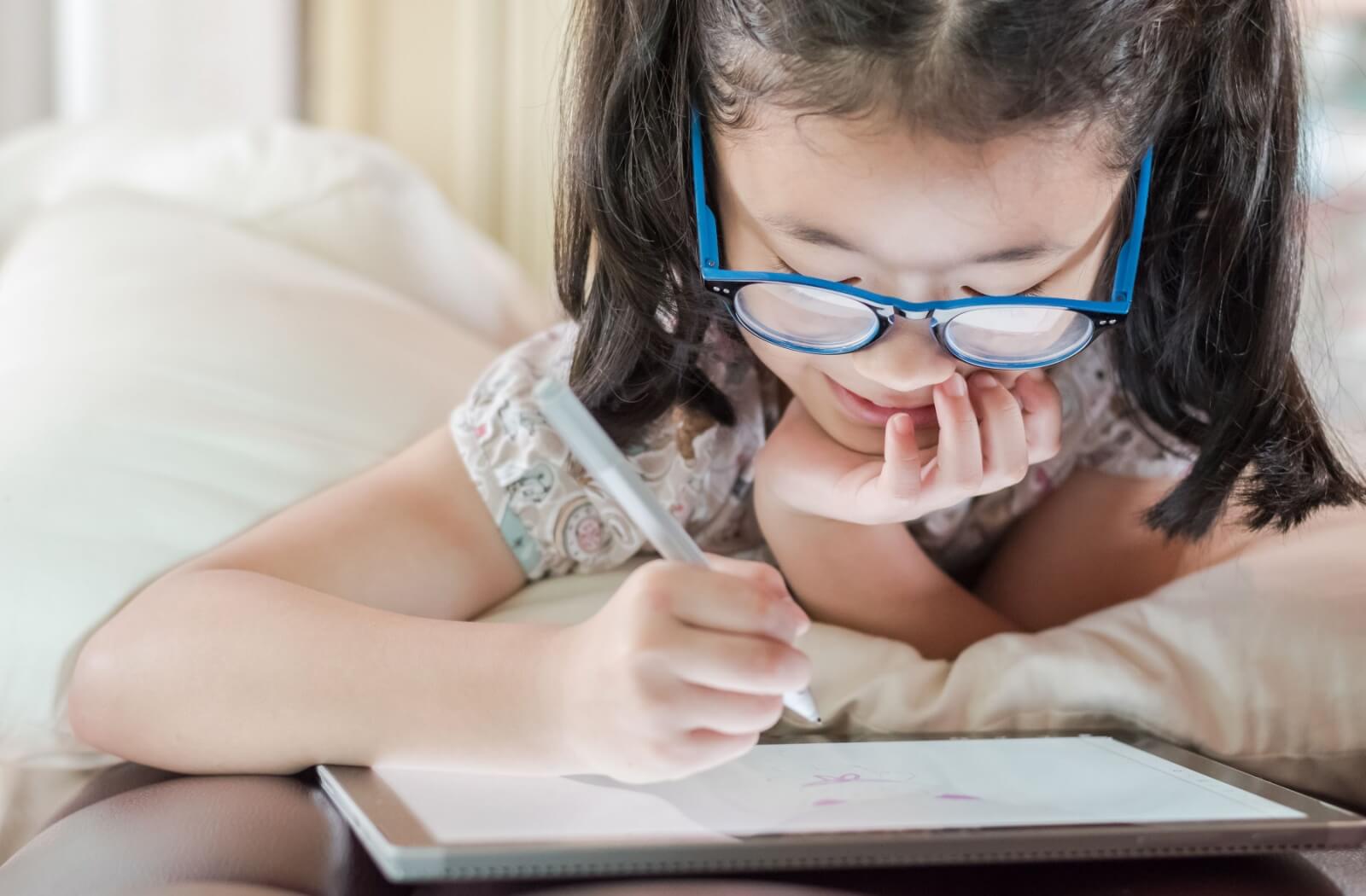 A school-age child wearing computer eyeglasses draws on a digital tablet while lying in bed