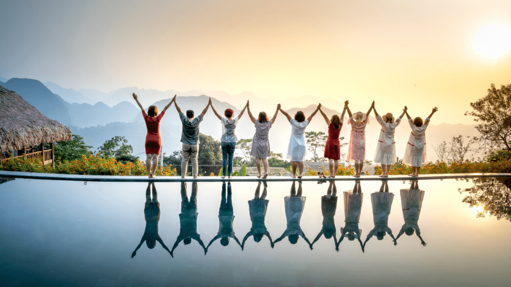 A diverse group of people stands hand in hand by an infinity pool, arms raised toward the sunset, symbolizing connection, empowerment, and shared growth.